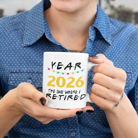 A person in a blue polka-dot shirt holds a white ceramic mug that reads 'YEAR 2026 THE ONE WHERE I RETIRED' in black and yellow text with small colorful flag accents.