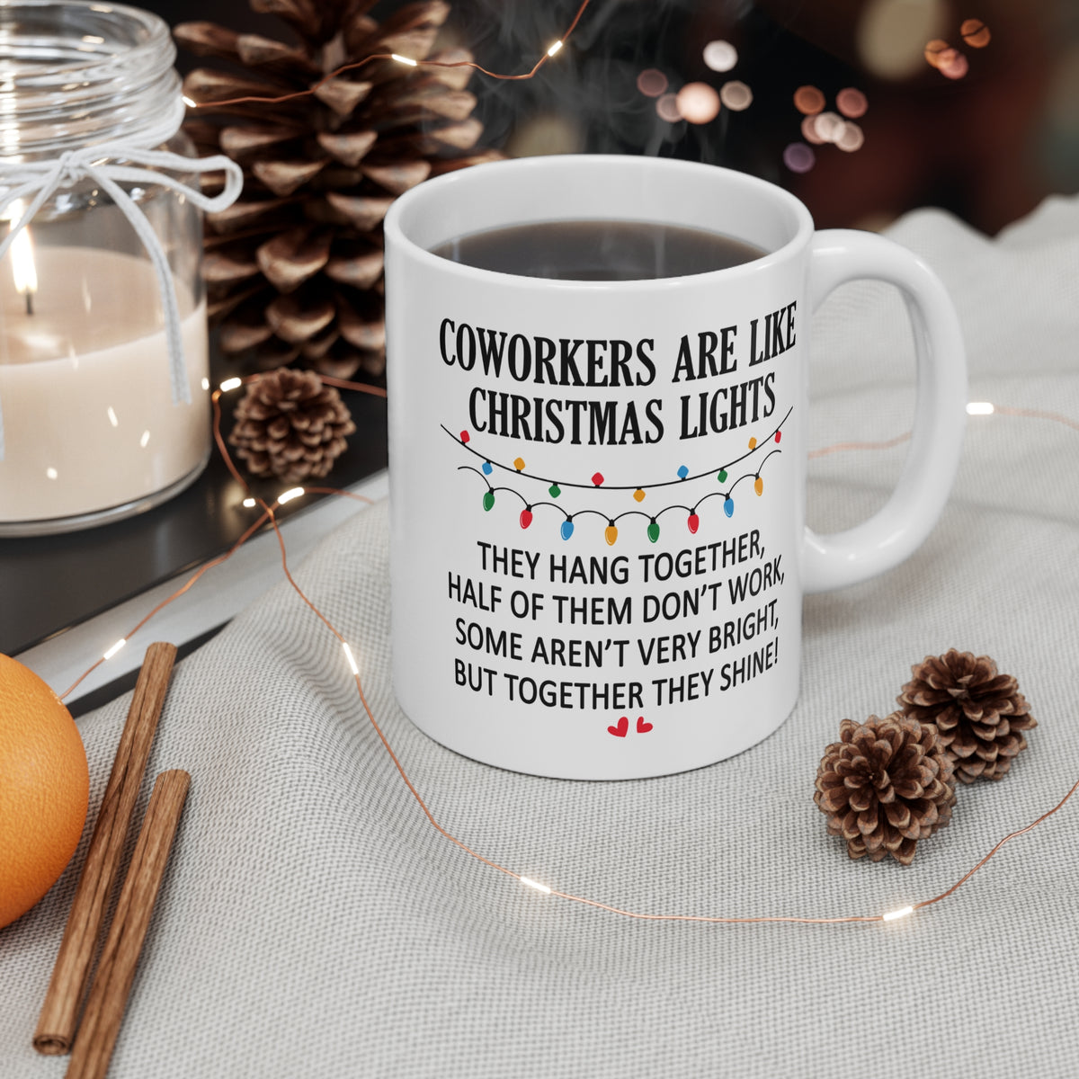 A white ceramic mug with a humorous Christmas-themed quote printed on it, styled on a light fabric surface with pinecones, fairy lights, cinnamon sticks, and a candle in the background.