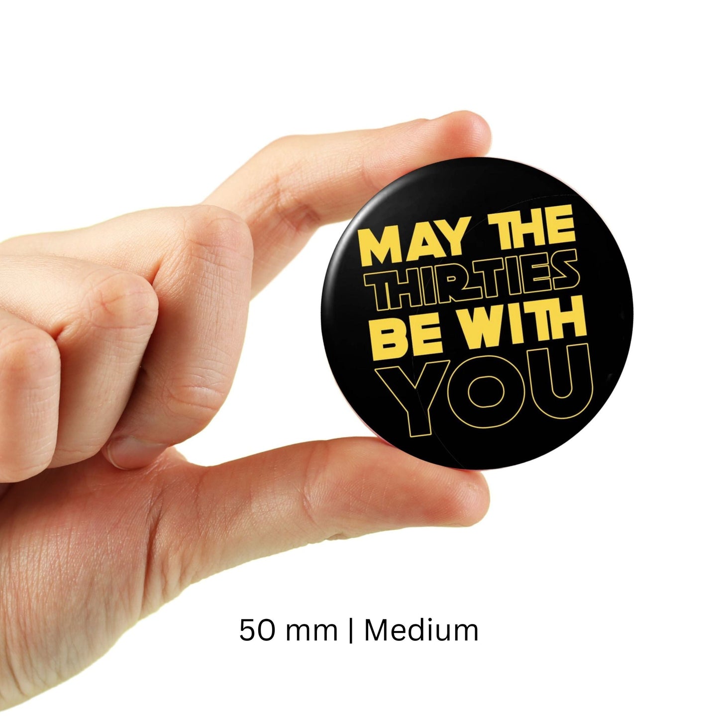 Hand holding a black button badge with yellow text 'May The Thirties Be With You', 50 mm medium size, birthday accessory.