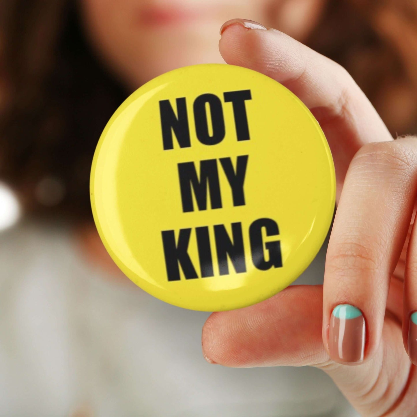 Close-up of a hand holding a yellow 'Not My King' anti-monarchy pin badge with bold black text, symbolizing political activism.