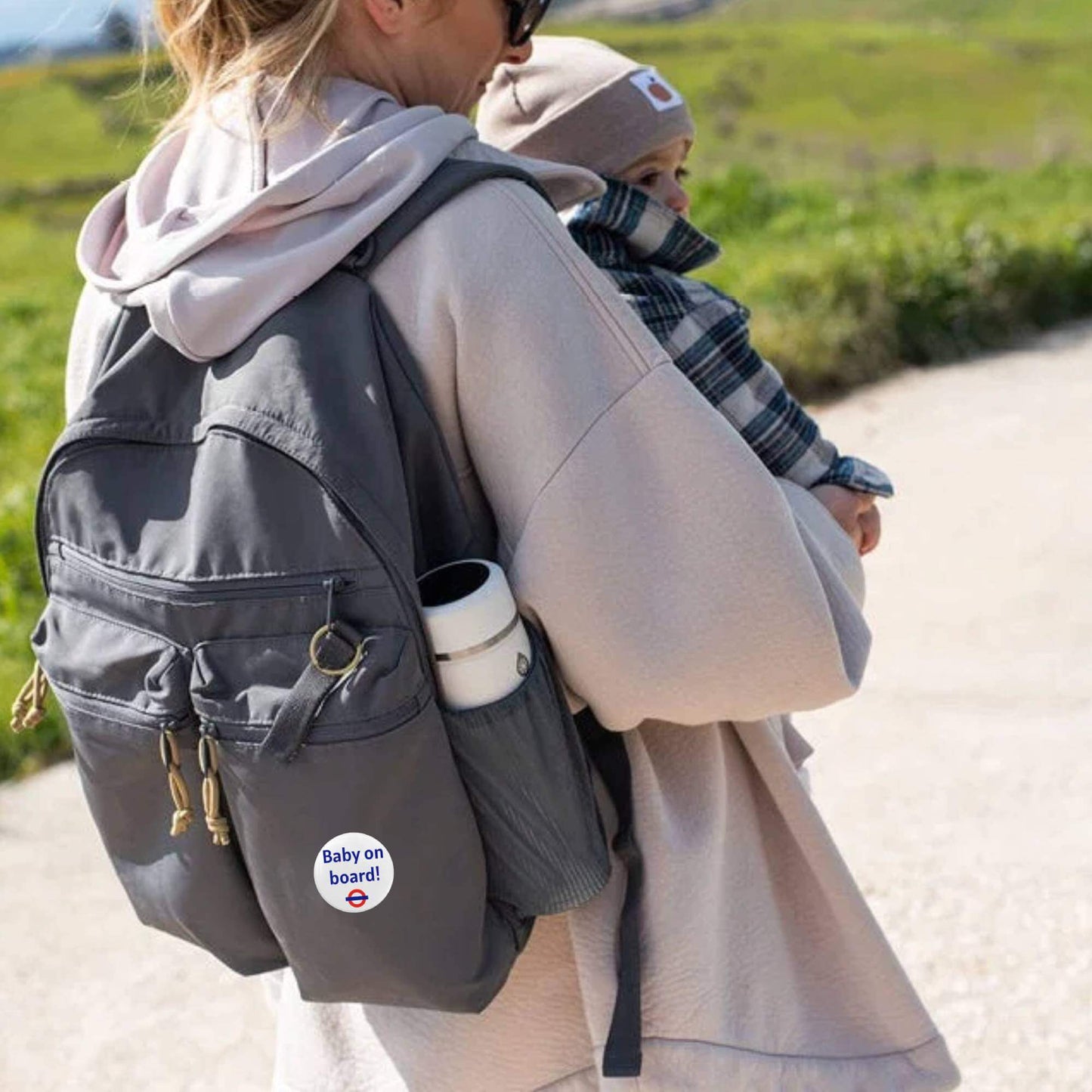 A woman carrying a baby and a backpack adorned with the EMU Works Baby On Board Pin, symbolizing excitement during pregnancy.