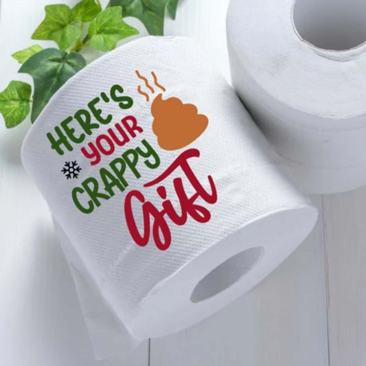 Close-up of a humorous bathroom decor toilet roll stating "Here's Your Crappy Gift" with a whimsical design and green and red lettering.
