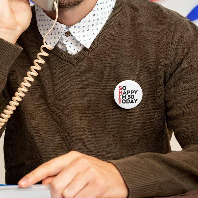 Man talking on the phone wearing a 50th birthday badge that says 'SO HAPPY I'M 50 TODAY'.
