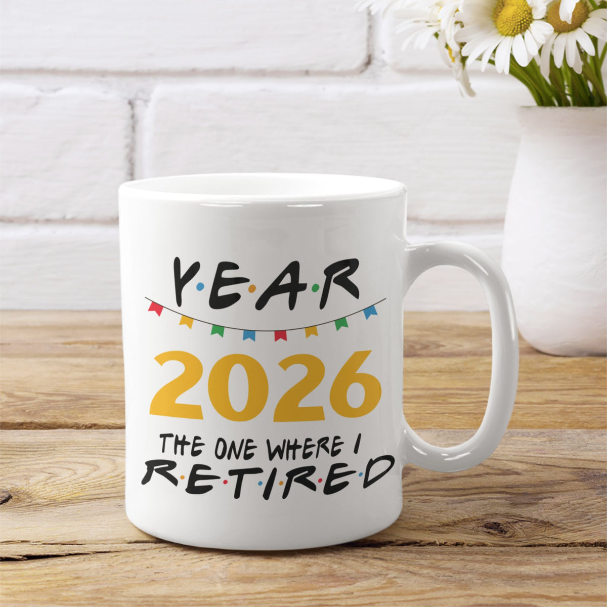 A white ceramic mug with a 'Year 2026 The One Where I Retired' graphic, displayed on a wooden table with a white brick wall and flowers in the background.