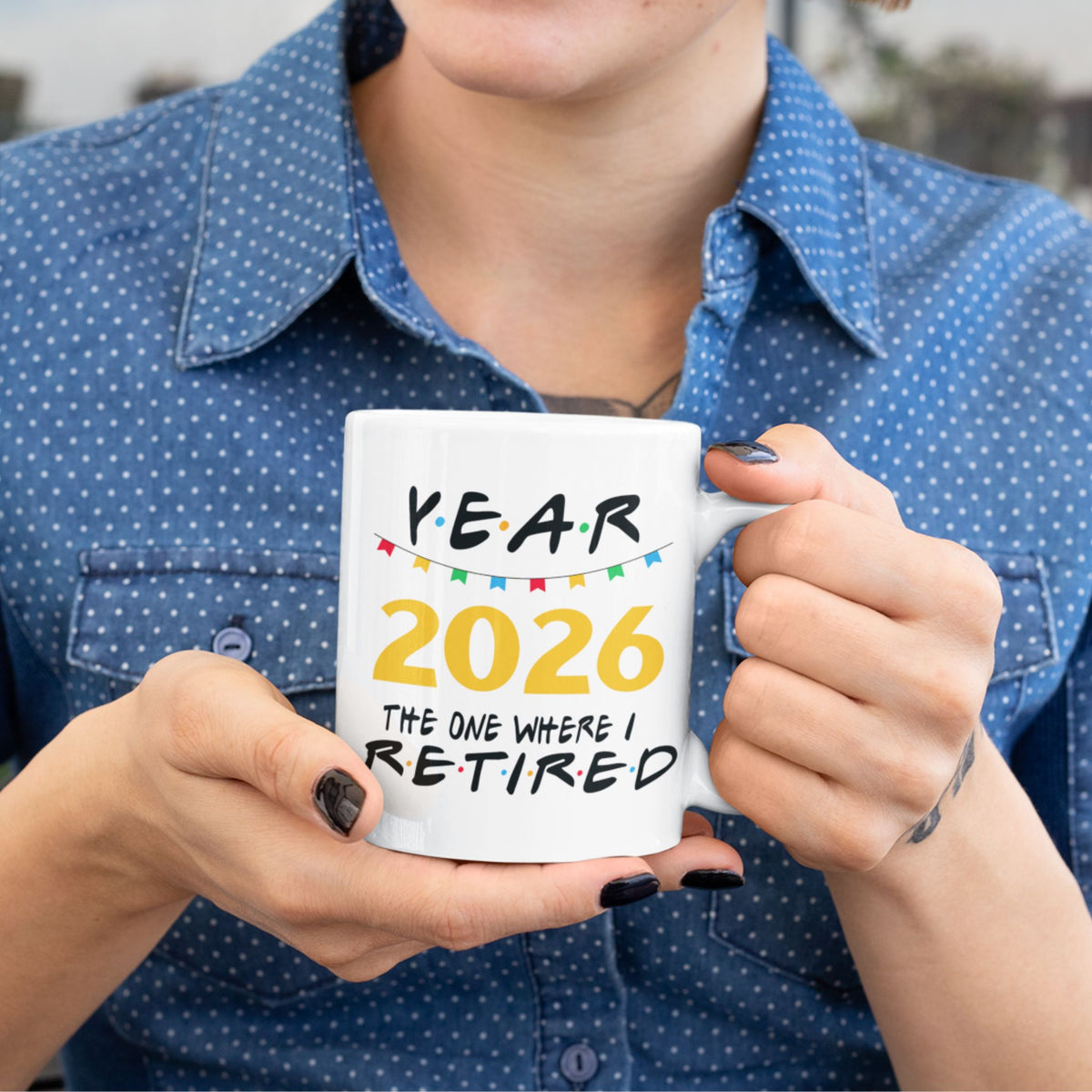 A person in a blue polka-dot shirt holds a white ceramic mug that reads 'YEAR 2026 THE ONE WHERE I RETIRED' in black and yellow text with small colorful flag accents.