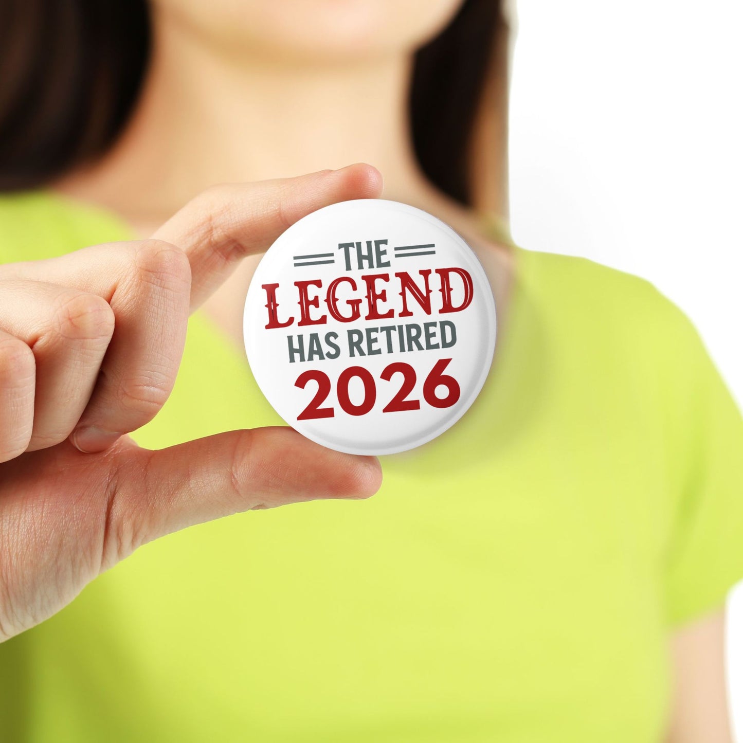 Person holding a button with 'The Legend Has Retired 2026' text against a white background