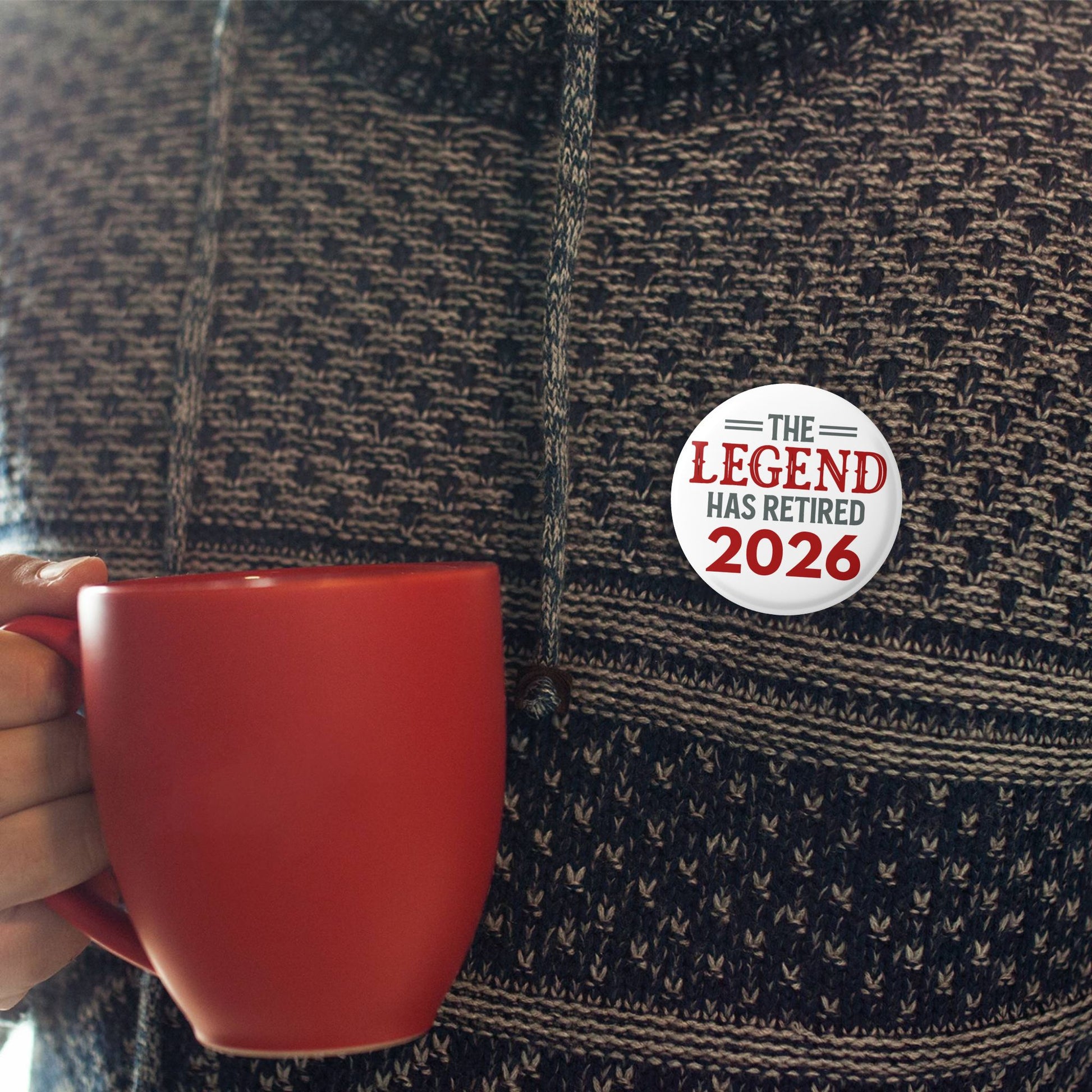 Person holding a red mug with a 'The Legend Has Retired 2026' button on a textured shirt.
