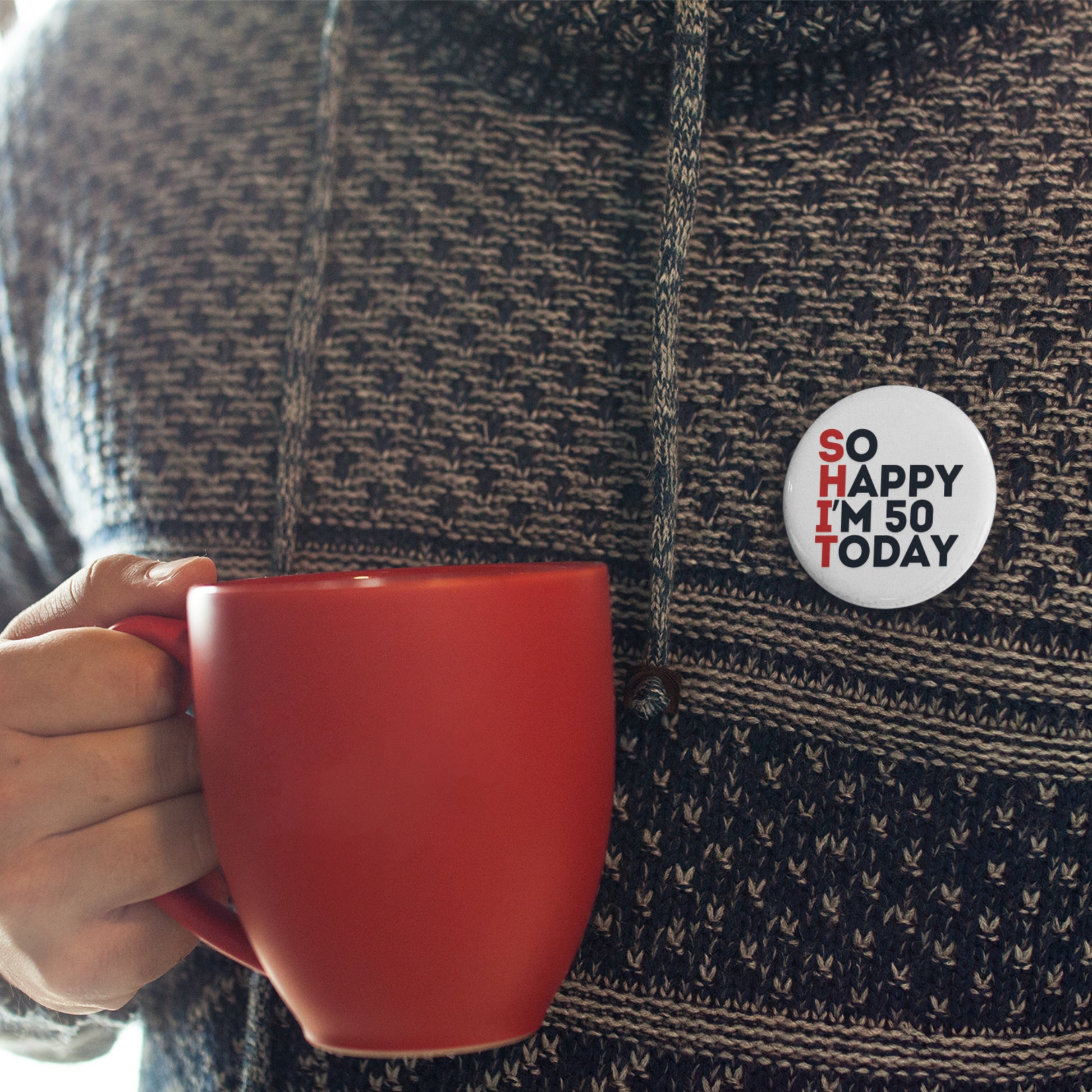 A close-up of a man in a patterned sweater holding a red mug and displaying a 50th birthday badge that says 'SO HAPPY I'M 50 TODAY'.
