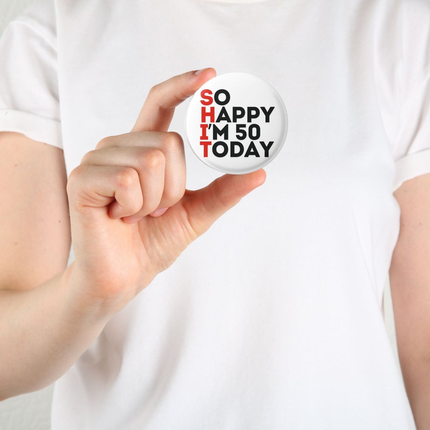 A person holding a 50th birthday badge that states 'SO HAPPY I'M 50 TODAY' against a plain white background.